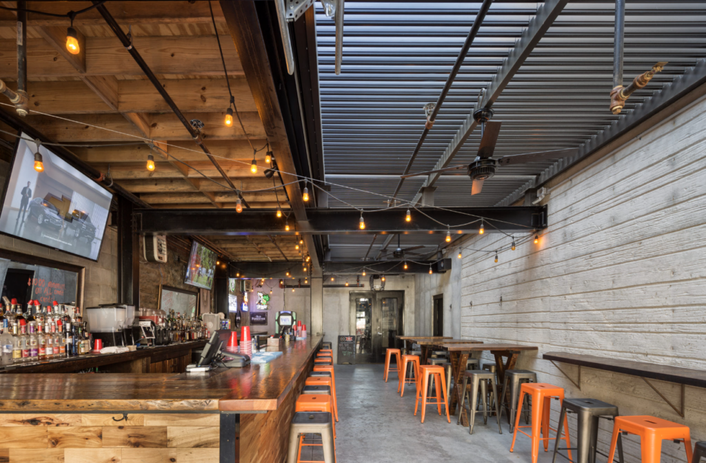 Industrial-style bar interior with Edison string lights, wood countertop, and orange metal stools.