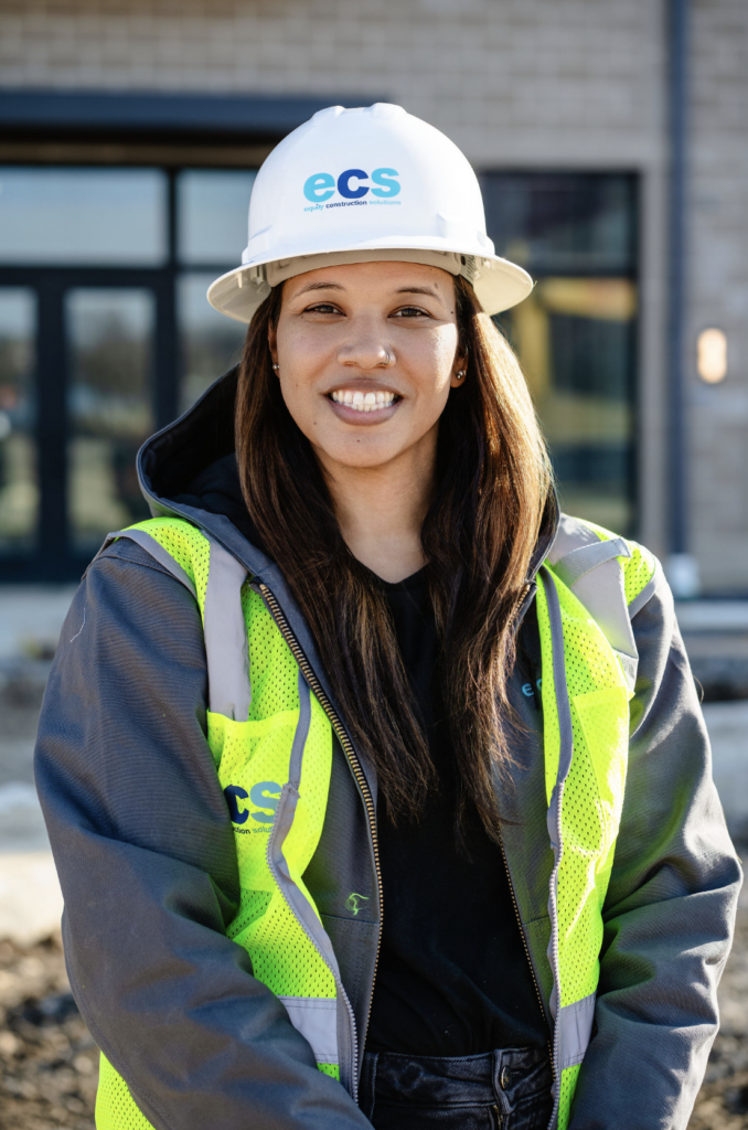 Smiling woman in an ECS hard hat and high-visibility vest at a construction site.